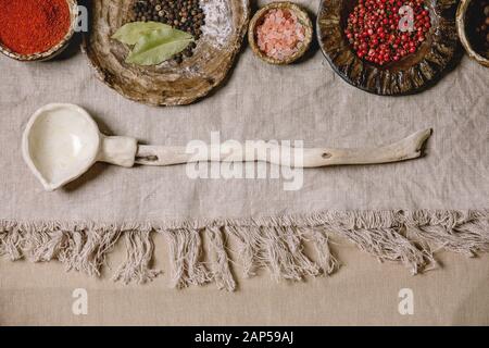 Variety of different handmade ceramic plates and bowls in row with seasonings and spices, and ceramic ladle over grey linen cloth as background. Flat Stock Photo