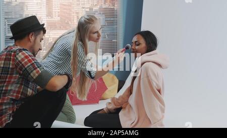 Close-up of make-up artist applying makeup on beautiful black model sitting on the floor and posing. Photographer taking photos on professional camera. Stock Photo