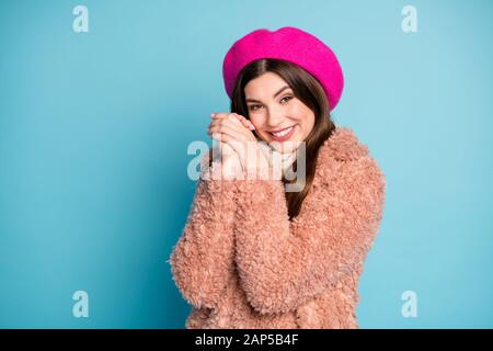 Portrait of glad dreamy girl in bright beret enjoy winter cold weather feel grateful hold fists near face wear fur pastel overcoat isolated over blue Stock Photo