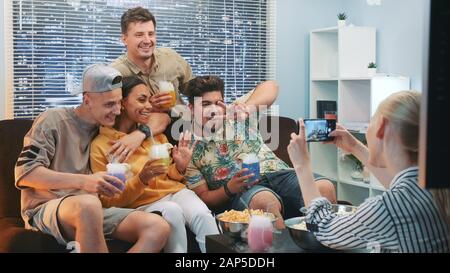 Close up of girl making photo of friends sitting on sofa with cocktails by smartphone. The group of people cheers, smiling and posing on camera. Stock Photo