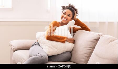 African American Woman Sitting On Sofa Embracing Pillow Indoor, Panorama Stock Photo