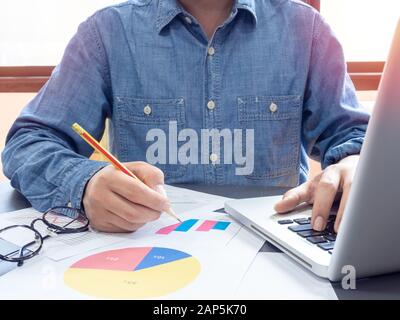 Man wearing blue shirt writing on paper and working with laptop computer on table near the window in office. Stock Photo
