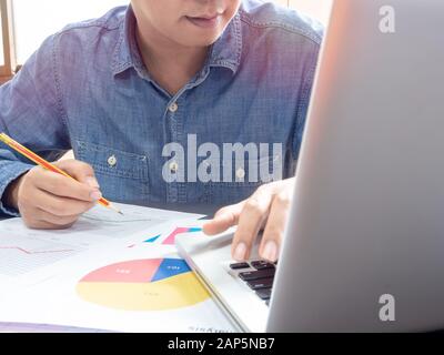 Asian businessman wearing blue shirt holding pencil and working with laptop computer on table near the window in office. Stock Photo