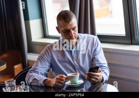 Cute elegant businessman reading a message on a smartphone in a restaurant on a table and drinking coffee. A man wears a shirt Stock Photo