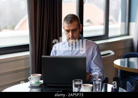 Cute elegant businessman working on a laptop in a restaurant on a table and drinking coffee. A man wears a shirt and sitting next to a big window Stock Photo