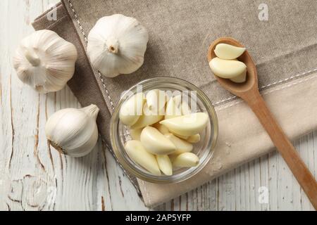 Glass bowl of fresh garlic bulbs, spoon of slices, napkin on white wooden background, closeup. Top view Stock Photo