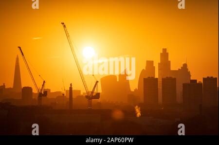 The sun sets behind tower cranes and the London skyline, including the Shard (left) and skyscrapers in the city financial district of London. Stock Photo