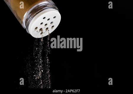 Black ground pepper falls out of the pepper shaker on a black isolated background. The seasoning process stop in time. Glass salt shaker with white st Stock Photo