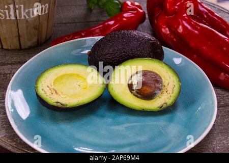 Close up of two isolated avocados on blue china dish and round wood table. Blurred basil pot and red bell pepper background. (German word Basilikum me Stock Photo