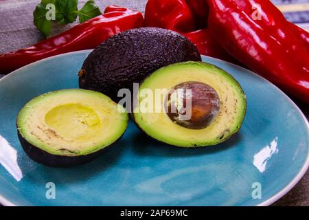 Close up of two isolated avocados on blue china dish and round wood table. Blurred basil pot and red bell pepper background. (German word Basilikum me Stock Photo