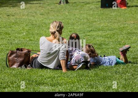 Rear view of a mother with brown hand bag relaxing with her two daughters on green grass while enjoying sunny day at park  Stock Photo