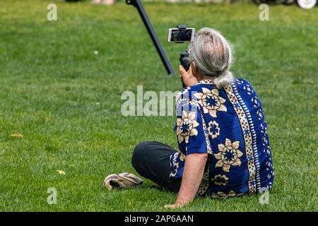 Male blogger in blue shirt recording video with Smartphone and tripod while sitting on grassy field in park during vacation  Stock Photo