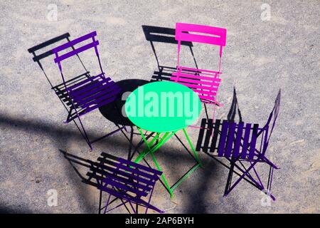 High angle view on green round table with four wood pink and purple folding chairs outdoor in bright sunlight throw shadows on grey gravel ground - Pr Stock Photo