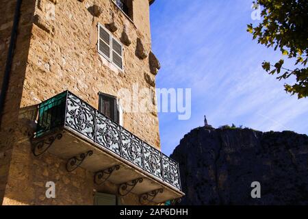 Low angle view on stone facade of typical French house and balcony with ancient ornate lattice work. Church on mountain top background - Castellane, F Stock Photo