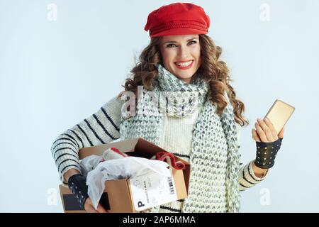 Portrait of happy young 40 years old woman with long brunette hair in sweater, scarf and red hat holding opened parcel and using smartphone app on win Stock Photo