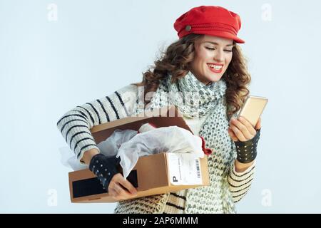 happy elegant woman with long brunette hair in sweater, scarf and red hat holding opened parcel and using smartphone app against winter light blue bac Stock Photo