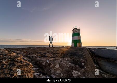 man with a backpack carrying a tripod looking out to sea by a lighthouse at sunset Stock Photo