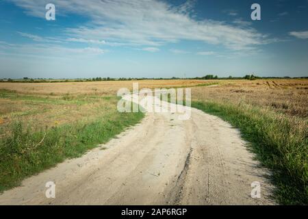 Winding dirt road through fields, horizon and white clouds on the blue sky Stock Photo