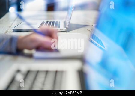 Closeup of hand woman writing working with a pen on a document, close to her computer laptop pc in her home office on her workplace, desktop career sc Stock Photo