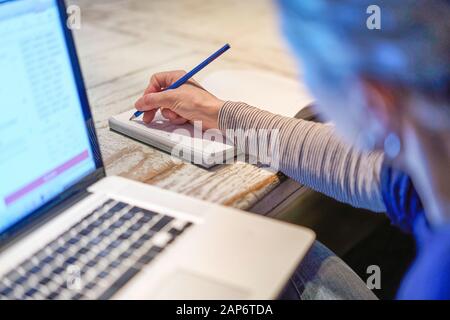 Closeup of hand arm face of woman writing working with a pen on a document, close to her computer laptop pc in her home office business family career Stock Photo