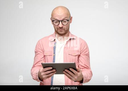 Bald man with glasses looks thoughtfully at the tablet that he is holding in hands. Stock Photo