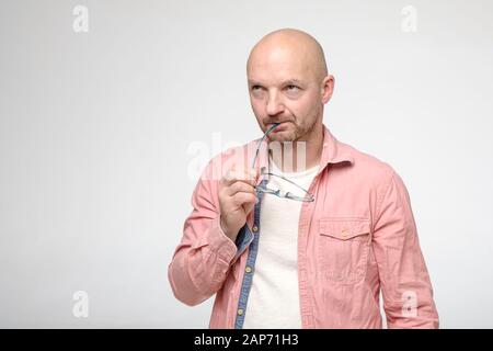 Upset, frowning bald man is trying to remember something, he holds glasses in hands and looks up thoughtfully. Copy space.  Stock Photo