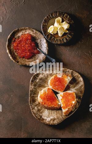 Red salmon caviar in bowl and on wheat bread, served with butter on ceramic plate over dark brown texture background. Flat lay, space Stock Photo