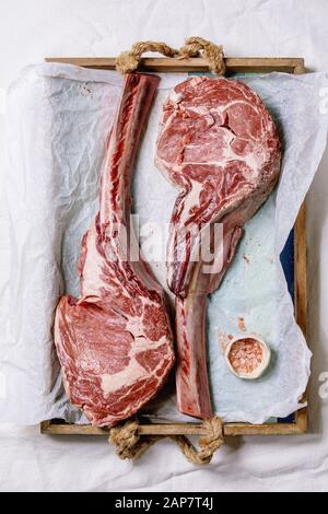 Raw uncooked black angus beef tomahawk steaks on bones served with pink salt on wooden tray with baking paper over white cloth as background. Top view Stock Photo