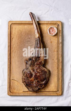 Grilled black angus beef tomahawk steak on bone served with pink salt on wooden cutting board over white cloth as background. Top view, space. Stock Photo