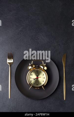 Interval fasting concept with a golden alarm clock on a black plate. Flat lay with copy space. Stock Photo