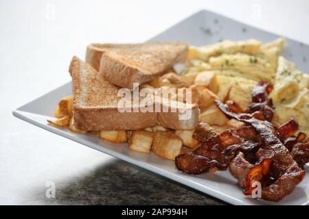 crispy well cooked pork bacon strips fried or grilled with hash browns and toast with tomato on plate in a close up view for breakfast lunch or dinner Stock Photo