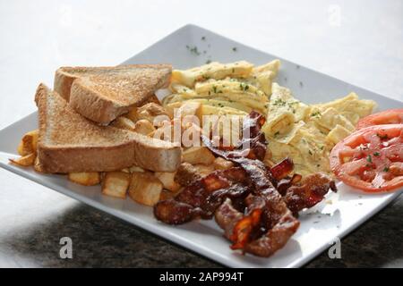 crispy well cooked pork bacon strips fried or grilled with hash browns and toast with tomato on plate in a close up view for breakfast lunch or dinner Stock Photo