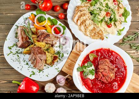 Different dishes on wooden table, top view. Vegetable beet soup, rack of lamb with fried potato, Grilled chicken Caesar salad. Stock Photo