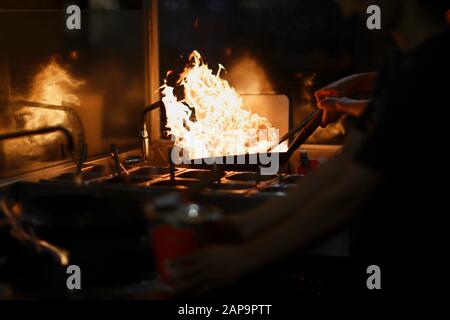 The cook in the kitchen holds a frying pan wok with an open flame and tries to extinguish it with water Stock Photo