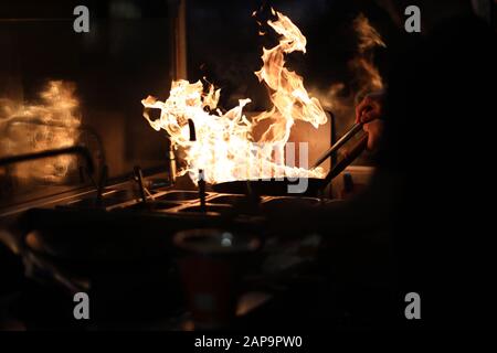 The cook in the kitchen holds a frying pan wok with an open flame and tries to extinguish it with water Stock Photo