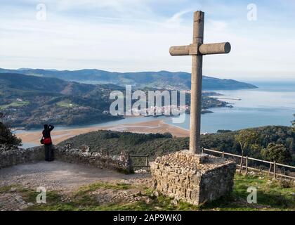 Hiker photographing Urdaibai estuary from San Pedro Atxarre viewpoint next to cross monolith with Mundaca and Machicacho cape (Basque Country, Spain) Stock Photo