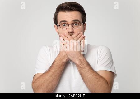 Head shot studio portrait millennial fearful man covering mouth. Stock Photo