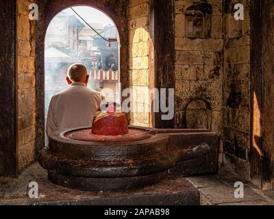 Local priest sitting behind a Shiva lingam in a small temple, watching a cremation ceremony at Pashupatinath temple Stock Photo