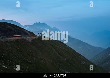A bus passes the Hotel Furka Blick on the Furka Pass mountain road at dusk in the Swiss Alps mountain top landscape of Realp, Uri, Switzerland EU Stock Photo