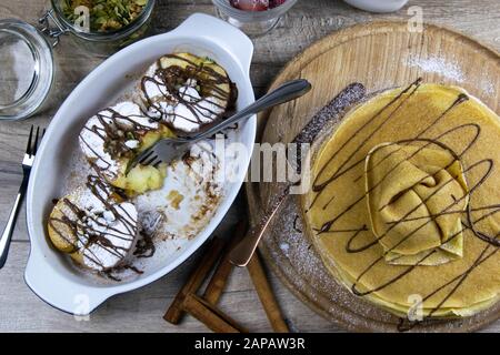 Caramelized apples with honey and cinnamon in a clay bowl and pancakes with butter and chocolate on a light wooden background. Top views Stock Photo