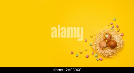 Golden brown eggs in a nest of multi-colored candy dragees on a yellow background with copyspace. Happy easter banner Stock Photo