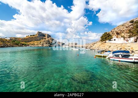 Lindos – view of St. Paul bay, motor boats anchored near orthodox church and acropolis of Lindos in background (Rhodes, Greece) Stock Photo