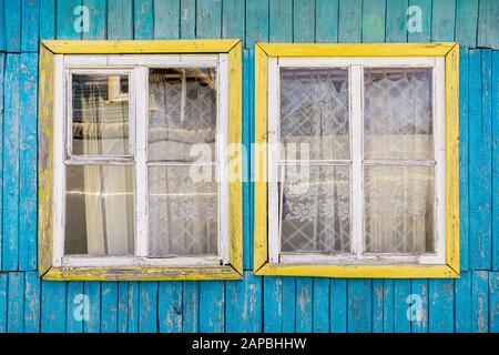 Fragment of wall of old wooden house with two windows with cracked paint on frames and holes between boards. Cornice with curtain behind glass. Rustic Stock Photo