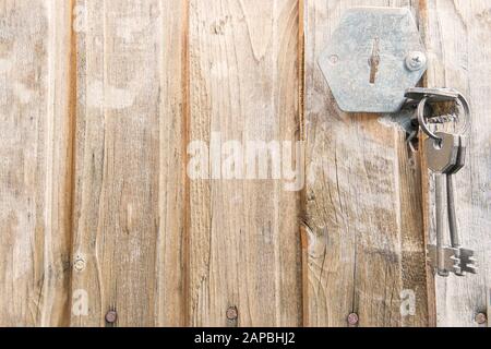 Old, cracked, weathered, wooden door with rusty nails and broken metal face. One iron key is inserted into keyhole and other two hang on ring. Stock Photo