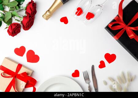 Valentines day dinner with table place setting, red tape, kraft gift box, glass for champagne, champagne bottle, roses, hearts, silverware on white ba Stock Photo