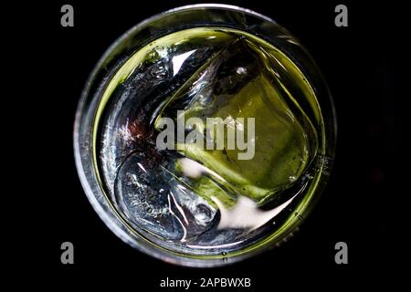 Gin tonic: View from above on isolated round cocktail glass rim with yellow green shimmering  ice cubes with lemon and cucumber. Black background. Stock Photo