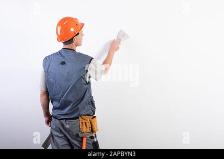 The builder smoothes the wall with a spatula. In work clothes and hard hat. On a light gray background. Stock Photo