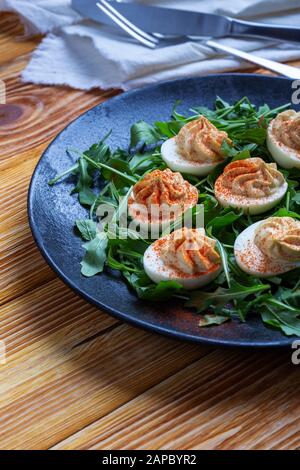 Deviled eggs with paprika on fresh arugula salad in black rural plate on wooden table, healthy vegetarian appetizer or snack close up Stock Photo