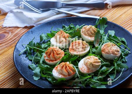 Deviled eggs with paprika on fresh arugula salad in black rural plate on wooden table, healthy vegetarian appetizer or snack close up Stock Photo