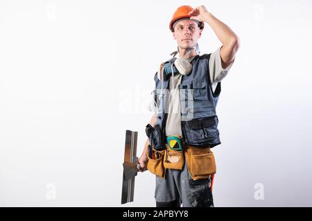 Builder with a spatula. Holding on to the helmet and looking forward. In work clothes and hard hat. On a light gray background. Stock Photo